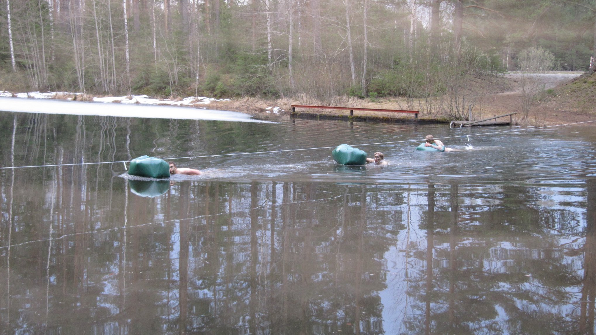 Three men swimming across a water with floating green bags. The water has some ice on it.