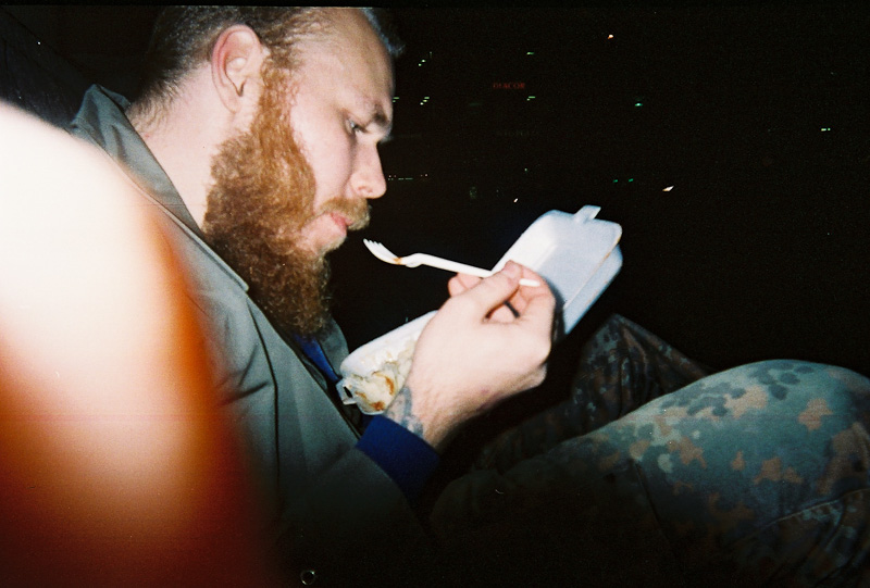 A bearded man eating from a styrofoam take away box with a plastic fork.