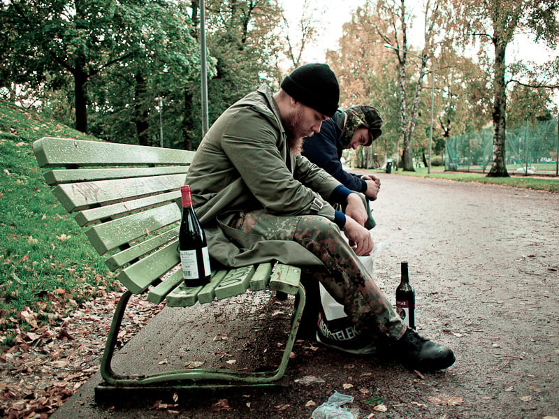 Two men sitting on a park bench bend forward and staring down in front of them. One wine bottle on the grounds and one on the bench.