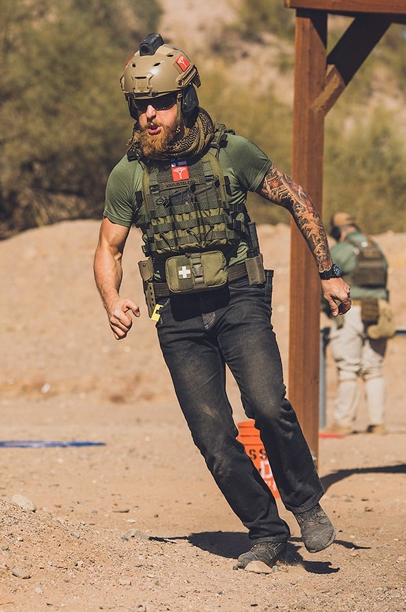 A fighter dressed in a helmet, body armor, battle belt, jeans and a t-shirt in a desert turnign to right appearing to be running.
