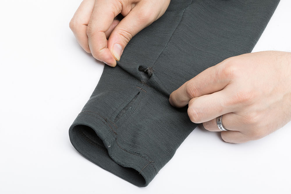 The end of a sleeve of a long sleeve shirt with the seam stretched with two hands above the thumb hole exposing a finger wide hole at the seam.