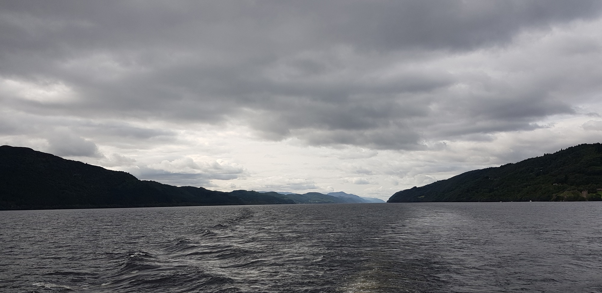 Stern waves of a boat on a lake, hills and grey sky.