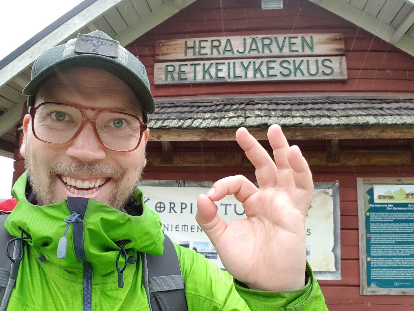 The hiker smiling widely and making an ok-sign with his fingers in front of Herajärven retkeilykeskus building.