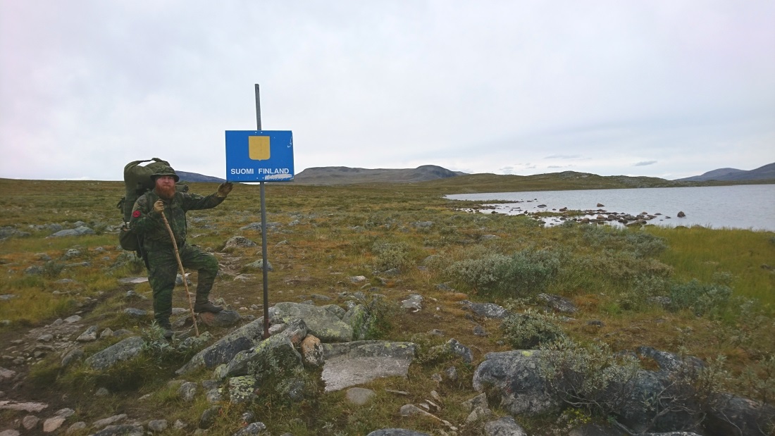 The bearded hiker posing next to a blue sign with the text SUOMI FINLAND next to a lake.