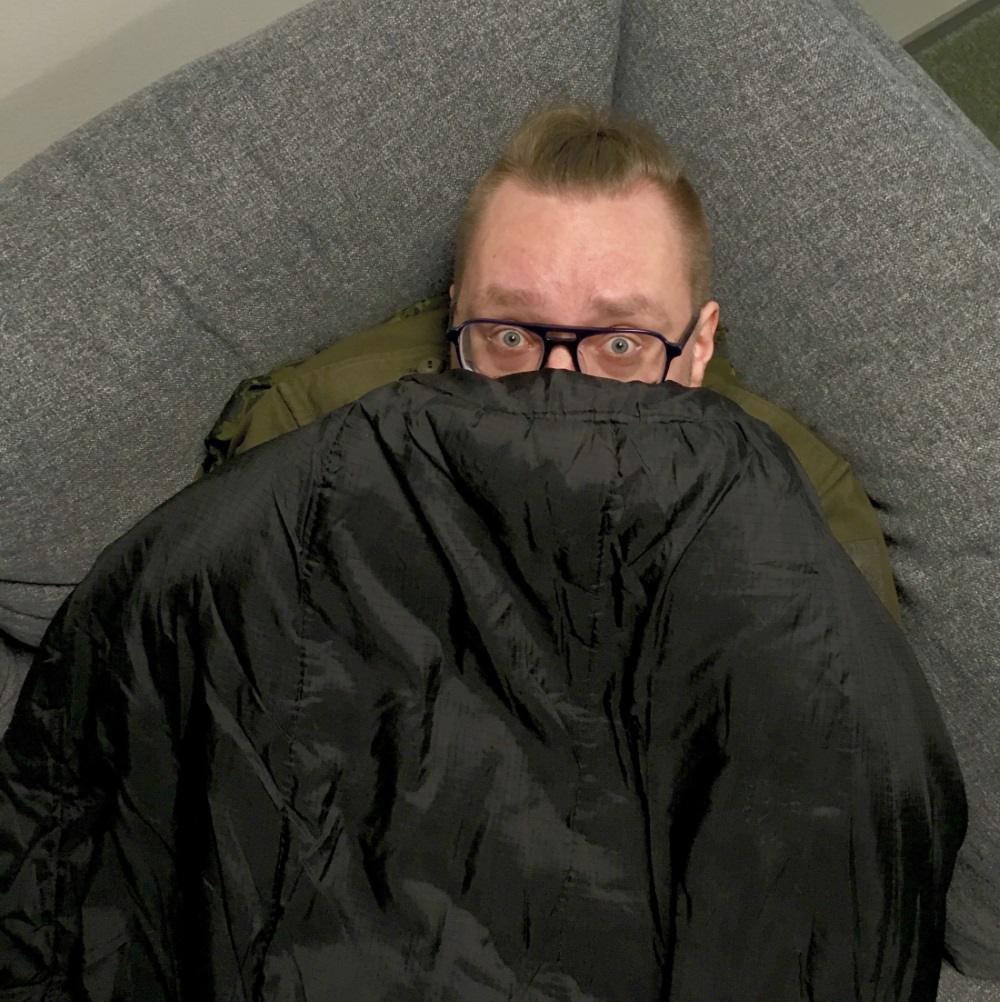A close-up of a person that appears to be lying on a couch under a sleeping bag that has been pulled up so that only the upper part of the face is visible. The person has a frightened expression on his face.