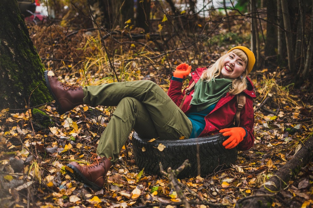 A woman in the fall woods on a car tire wearing brown boots, green pants, a turquoise jumper, a red jacket, a green scarf, orange gloves and a yellow beanie.