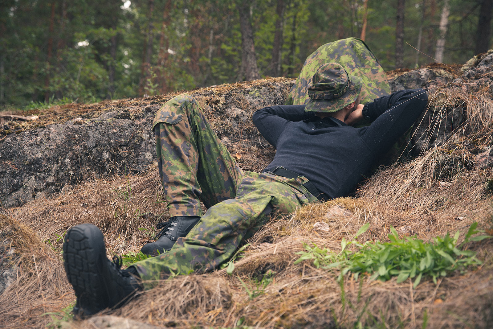 A man lying down on a grassy rock land with camo pants and hat and a black shirt and boots on.