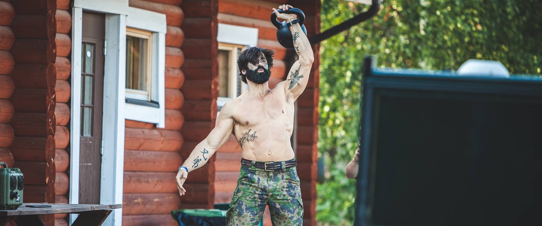 A muscular man without a shirt lifting a kettle ball in front of a red log house.