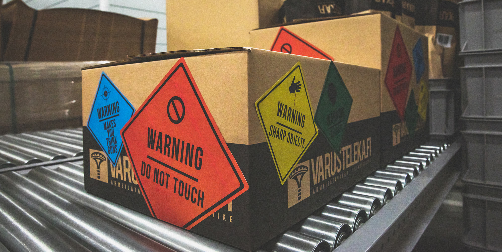 Varusteleka boxes on a conveyor belt with different colored warning stickers on them.