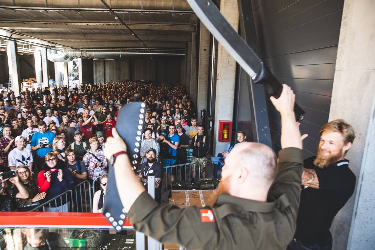 A large crowd has gathered into an industrial space with two men on a podium one of whom is holding up a rather large knife and its sheath and the other one watching him with a smile on his face.