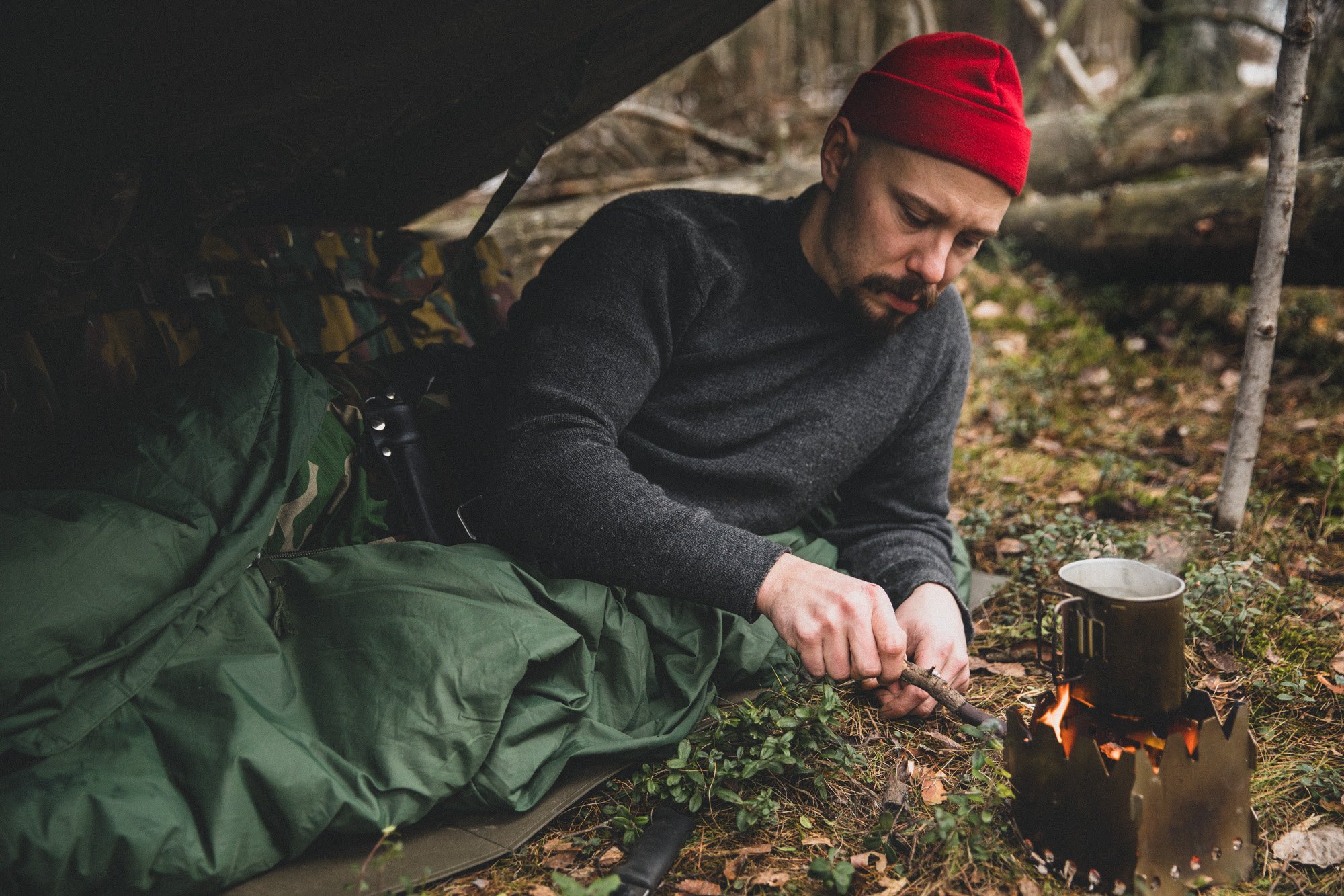 A man half in a sleeping bag lying in the forest under an awning feeding a stick into a twig stove.