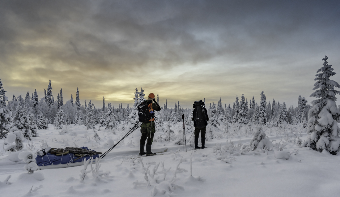 Kaksi metästäjää suksilla ihailee auringon kultaamaa taivasta talvisessa metsässä.