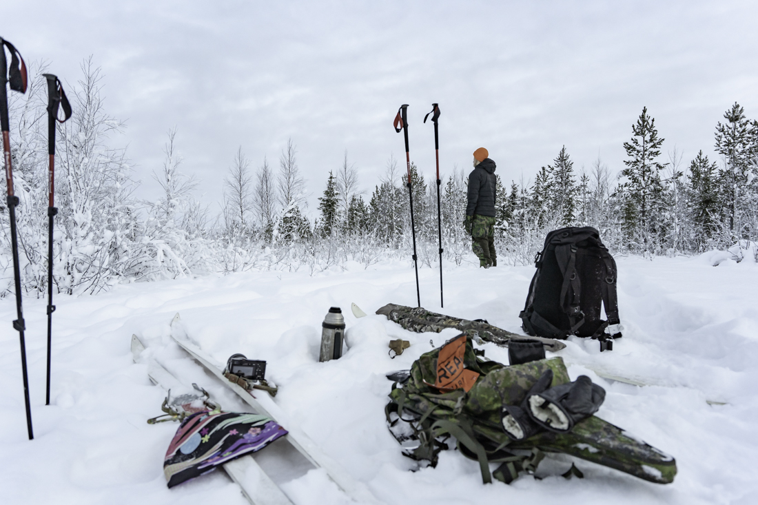 Taukopaikka metsän keskellä, varusteet maassa, metsästäjä tähyilee kaukaisuuteen.