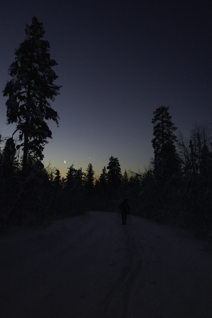 Hiihtäjä iltahämärässä, taivaalla kuunsirppi ja iltatähti.