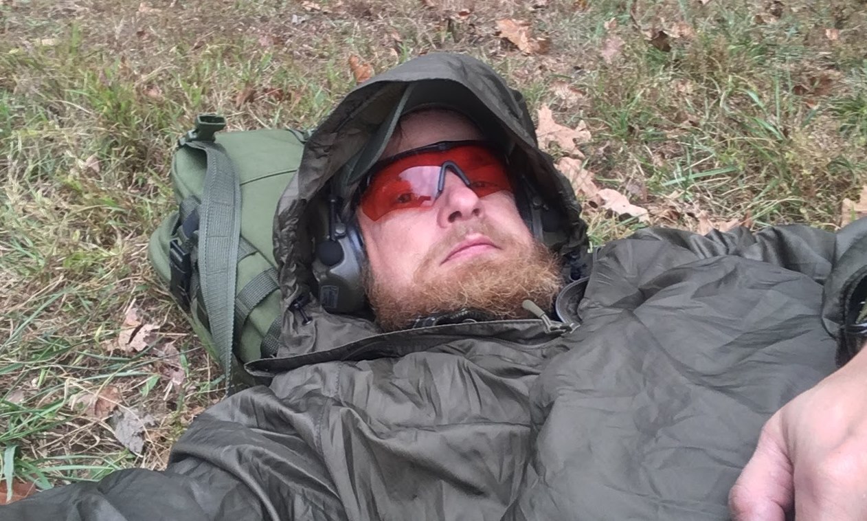 A man lying on the grass with a hood on his head and a backpack as his pillow.