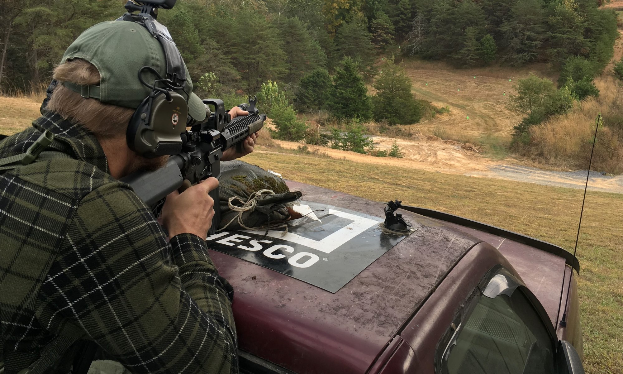 Man on a car on a hill shooting at green targets down below.