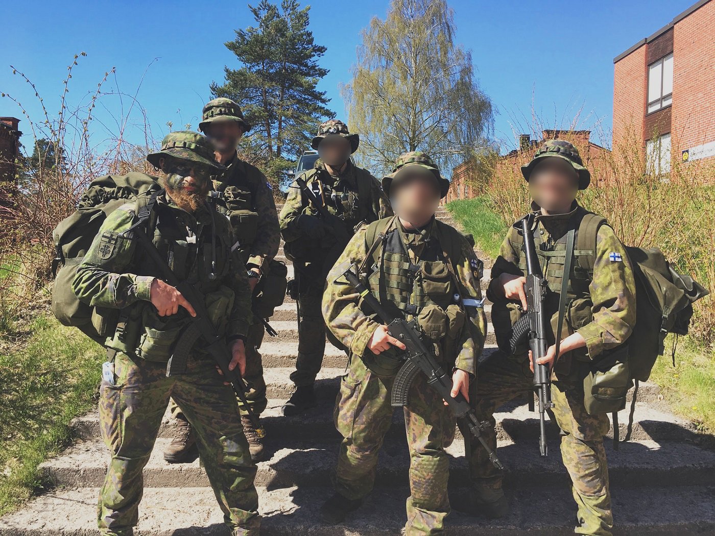 Five soldiers all geared up on stairs outside next to some brick buildings.