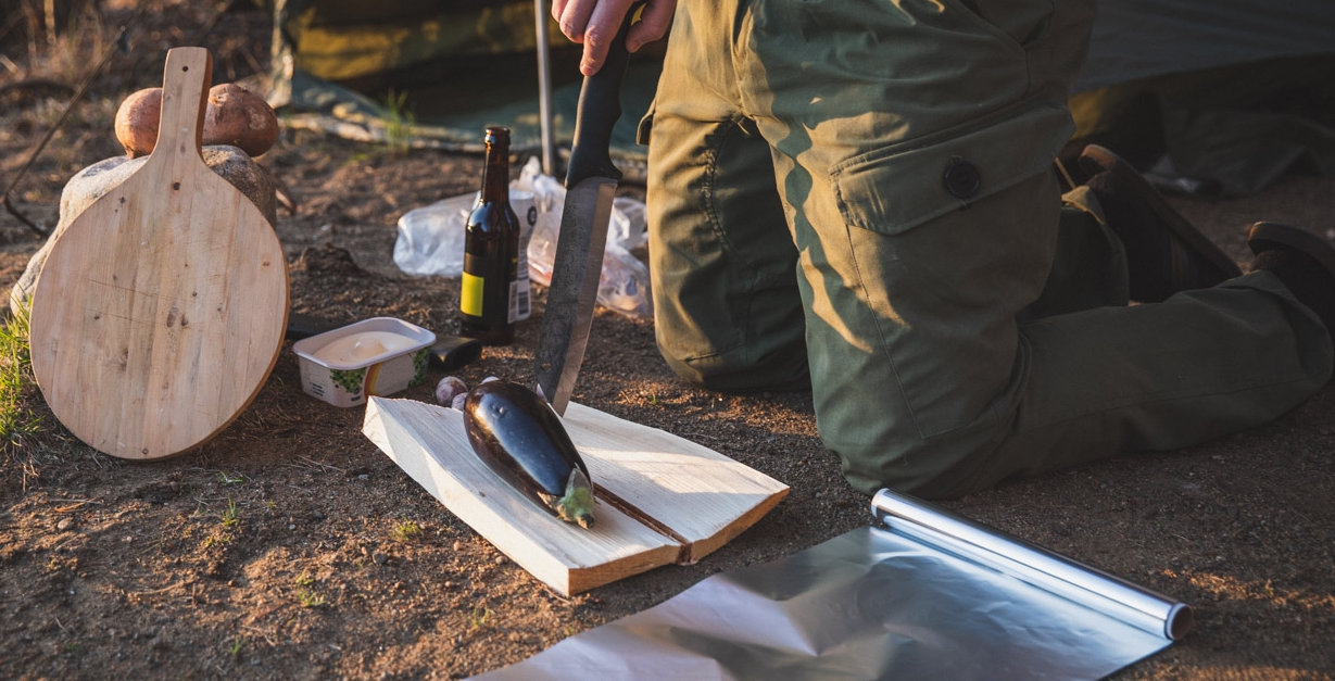 An eggplant on a chopping board made of log pieces on the ground with a man kneeling beside it and other cooking stuff around.