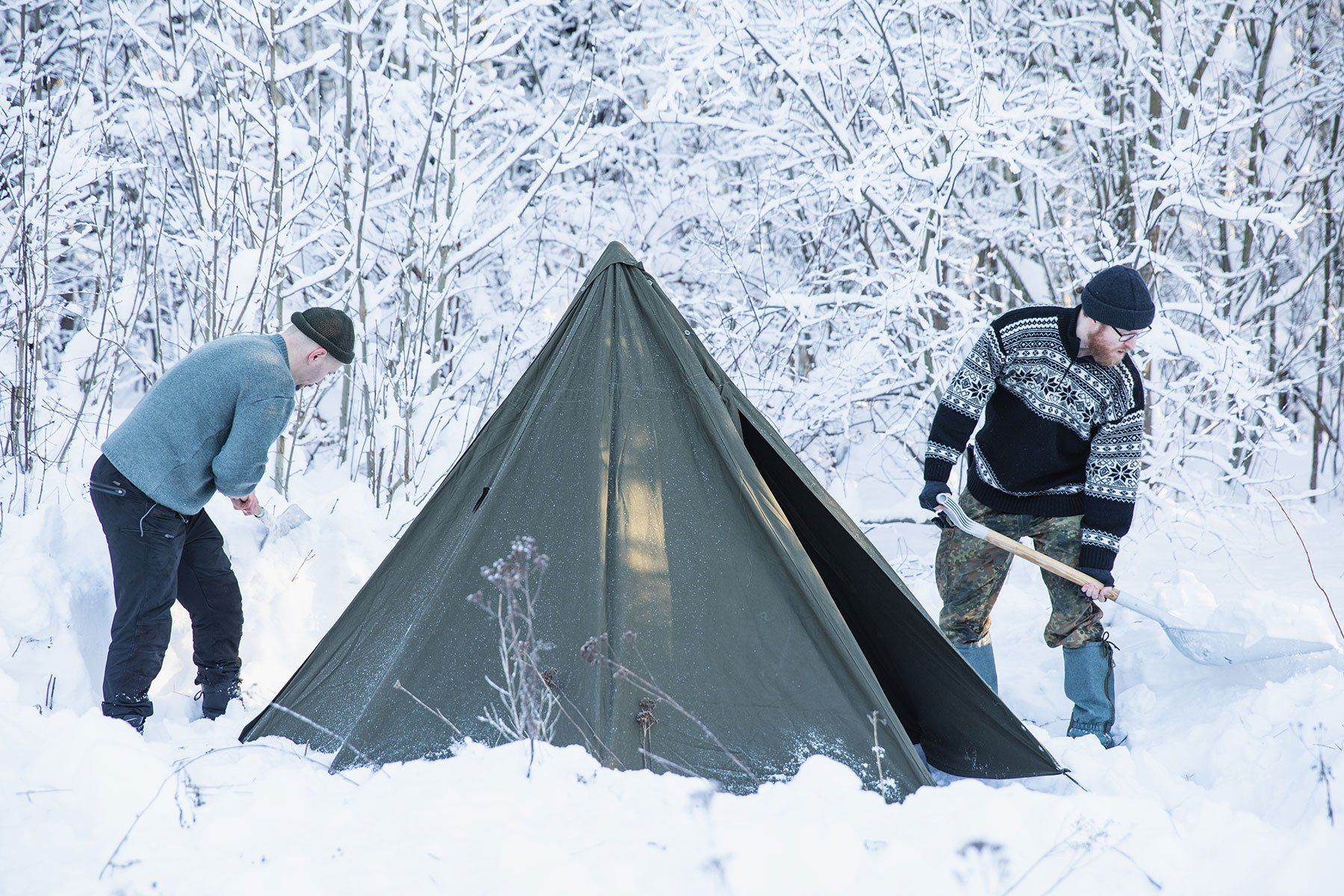 Two guys at a snowy forest edge on either side of a tent set up in the snow, shoveling snow from around the tent.