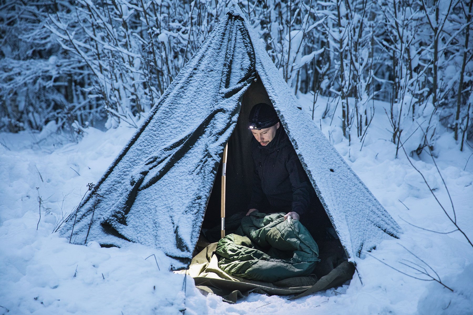 A person inside the tent in a wintry forest edge opening their sleeping bag.