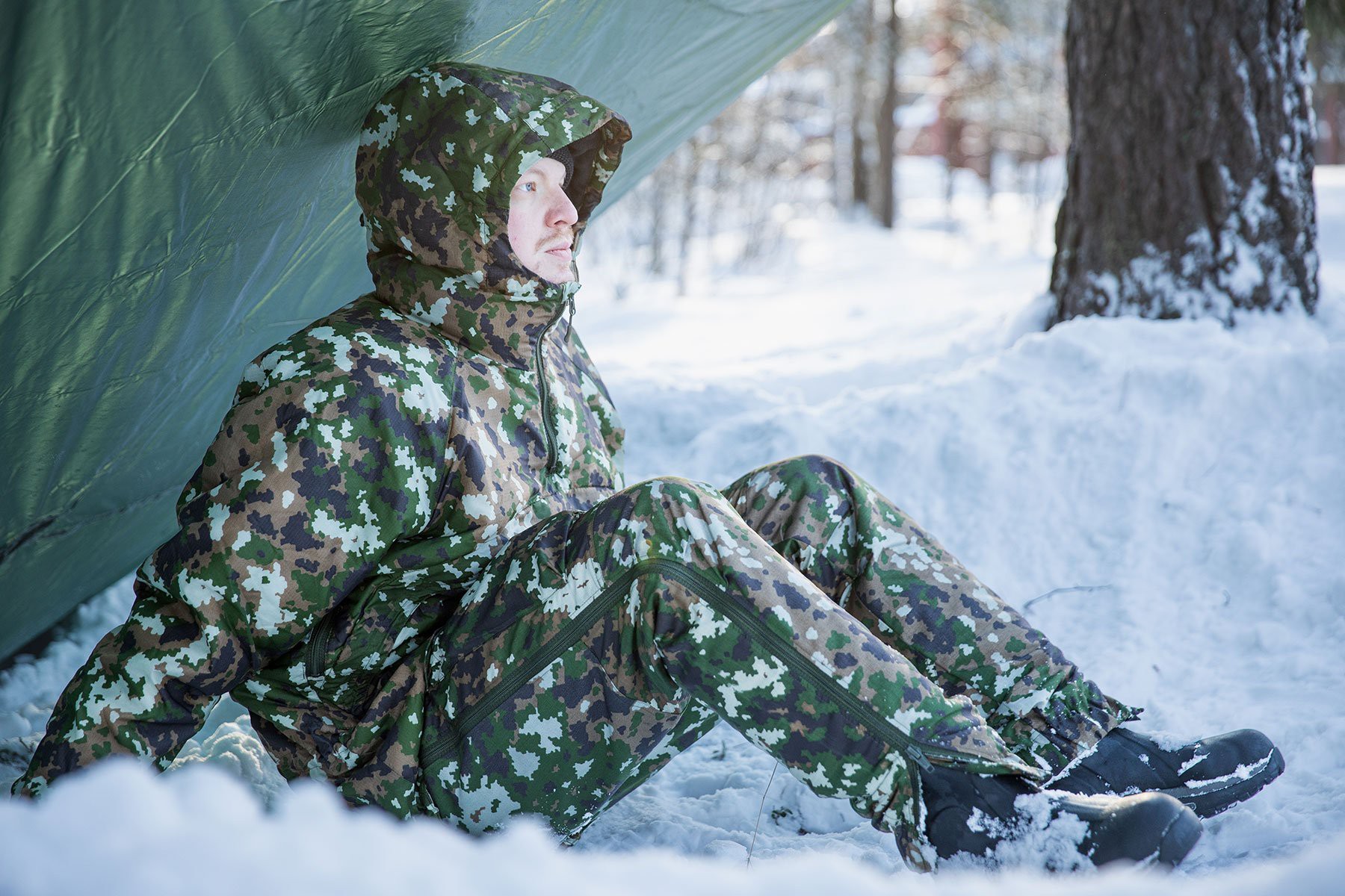 A person sitting under a tarp in snow, wearing a thermal suit in M05 camo.