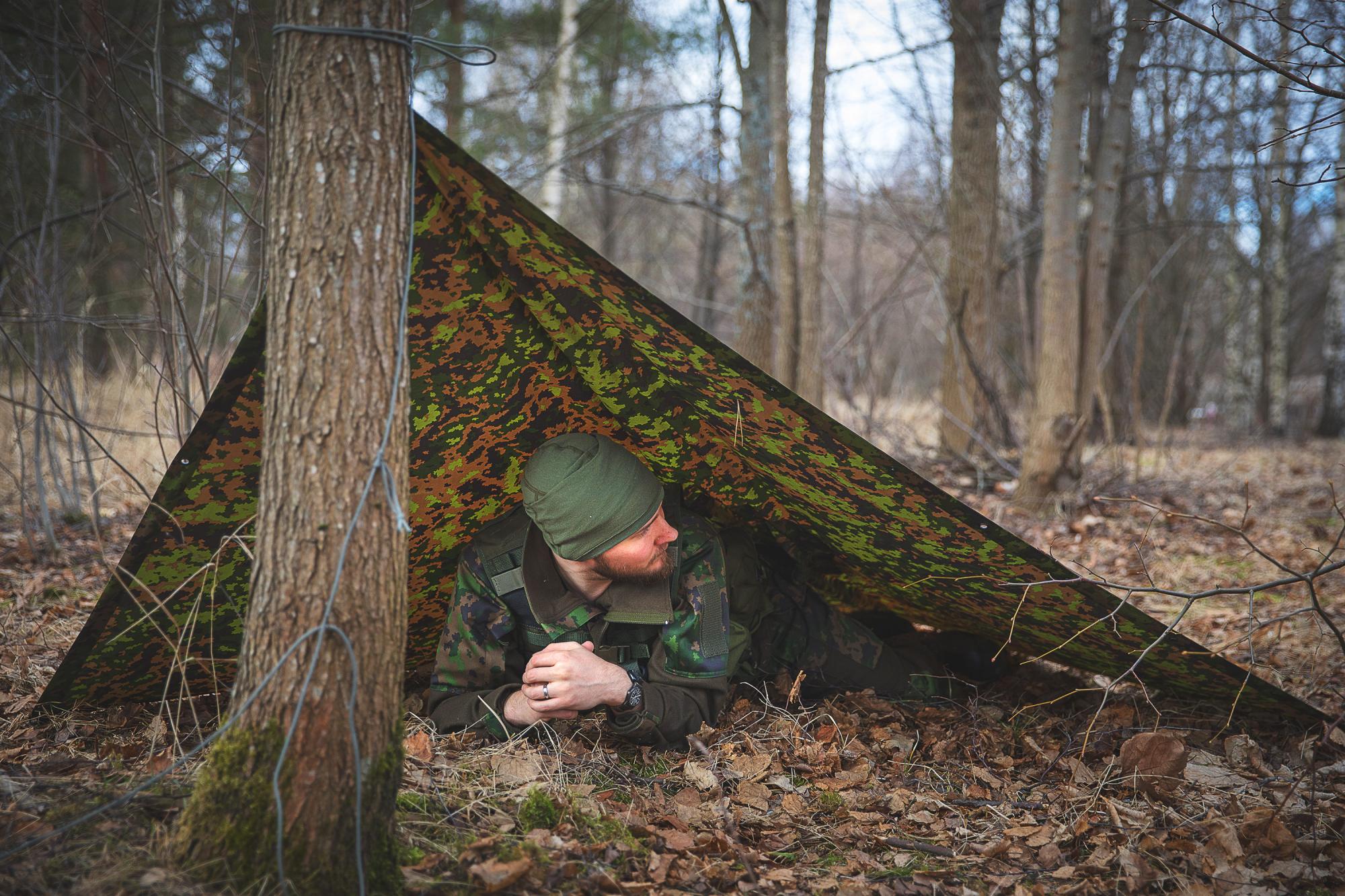 A man in a camo outfit lying down under a camo tarp 