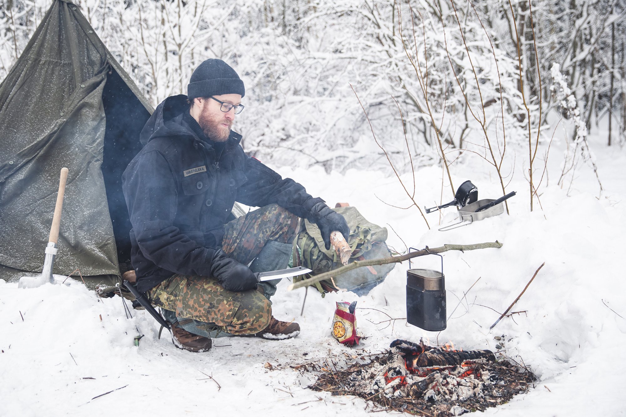 Bushcrafter cooks food in a mess tin on open fire in a wintery forest. 
