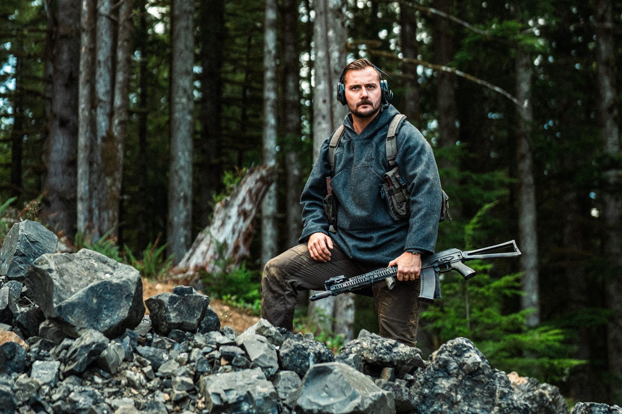 A man standing on a rock with Blanket Shirt on and a gun in his hand.