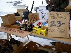 A sales table set up on snow, selling stuff like military surplus, patches, energy drinks and chocolate bars out of cardboard boxes.