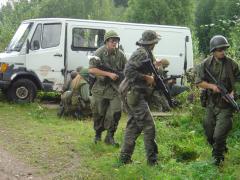 A rusty, old white van at the side of a dirt road with a group of people in military gear taking cover right next to it.