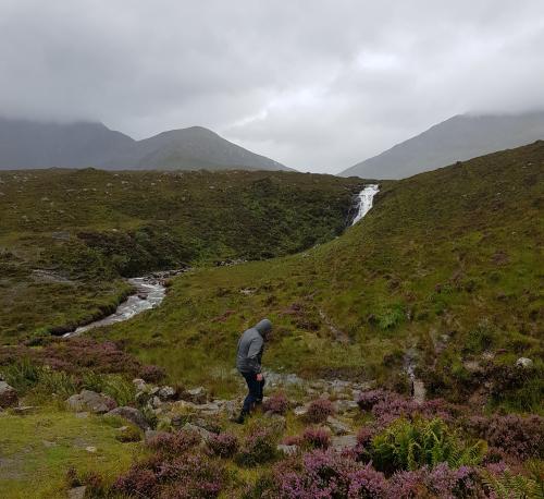 A scenery with a grassy and heathery field across which a creek flows. On the background grey clouds envelop hills. A person is making his way across the field.