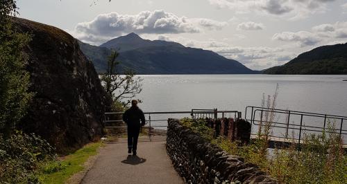 A mountainous lakeside scenery with a person walking towards the lake on a road leading onto a bridge.