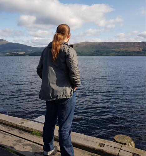 A long-haired man gazing out onto a lake and the hills on the other side of the lake.