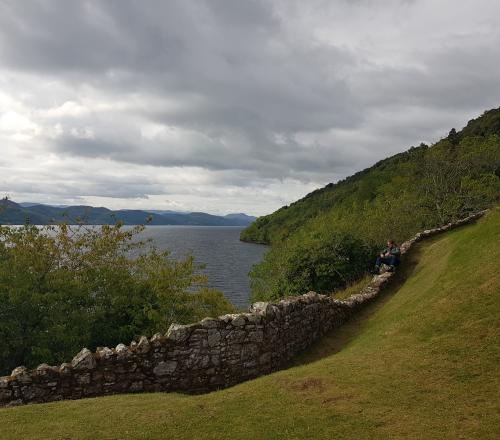 A man sitting on a stone wall on a green slope next to a lake.